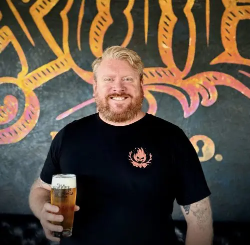 Matt wearing a black shirt while holding a beer in front of our logo wall.