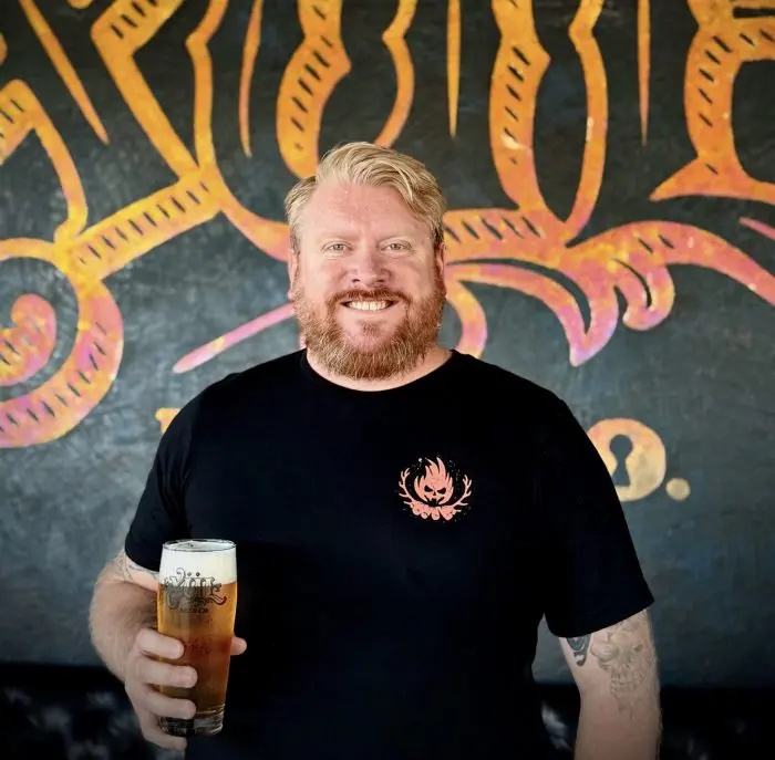 Matt wearing a black shirt while holding a beer in front of our logo wall.